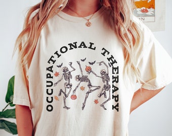 May include: Cream-colored t-shirt with the words "OCCUPATIONAL THERAPY" in a black arch. The shirt features a graphic of three dancing skeletons holding pumpkins and trick-or-treat buckets, with bats and leaves.