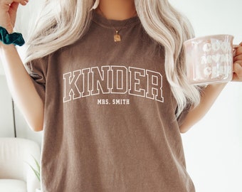 May include: A brown t-shirt with the text "Kinder" in white outlined letters and "Mrs. Smith" in smaller white letters below. The shirt is worn by a person with long blonde hair and a green scrunchie on their wrist.