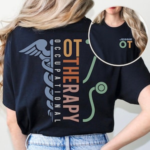 May include: Black t-shirt with a colorful graphic of a medical caduceus and the text "OT THERAPY OCCUPATIONAL". The shirt also has a small pocket on the left chest with the text "JESSIE OT" in white.