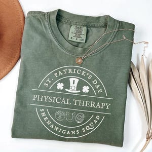 May include: A green t-shirt with a white circle design that says "St. Patrick's Day Physical Therapy Shenanigans Squad". The design includes a leprechaun hat, shamrocks, a pot of gold, a horseshoe, and a four-leaf clover.