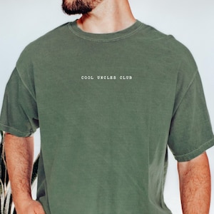 May include: Olive green t-shirt with the text "COOL UNCLES CLUB" in white. The shirt has a crew neck and short sleeves. The fabric appears to be a soft, washed cotton.