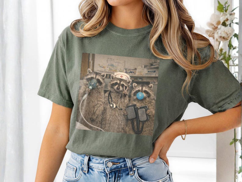 May include: Olive green t-shirt featuring a graphic of three raccoons in medical attire. One raccoon wears a nurse's cap, another has a blue bubble on its nose, and the third holds medical equipment. The image is a humorous take on healthcare.