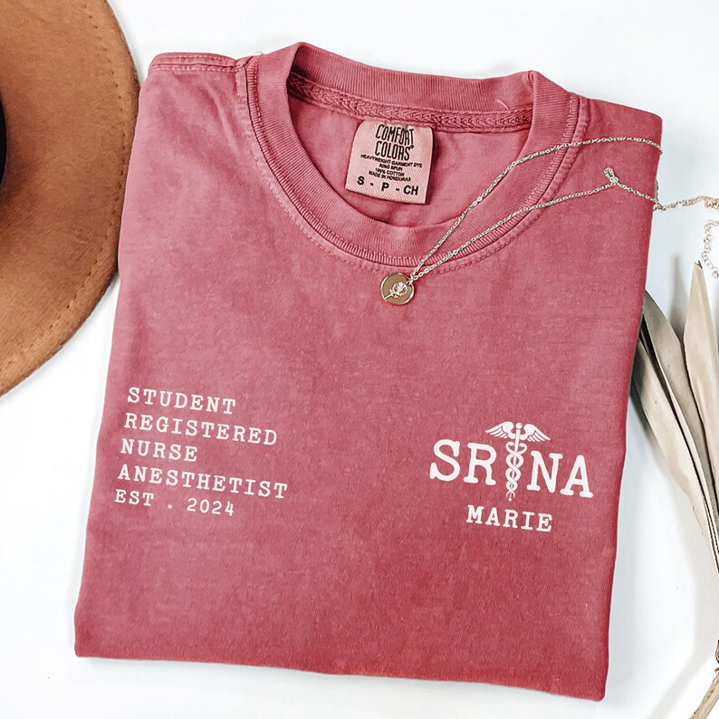 Może przedstawiać: Czerwony T-shirt z tekstem "STUDENT REGISTERED NURSE ANESTHETIST EST. 2024" po lewej stronie i tekstem "SRNA MARIE" z symbolem kaduceusza po prawej stronie.