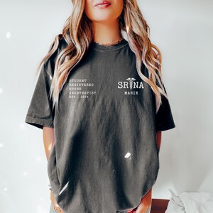 Może przedstawiać: Czarny T-shirt z białym tekstem, kt&oacute;ry brzmi "Student Registered Nurse Anesthetist  EST. 2024" i symbolem kaduceusza. Tekst "SRNA MARIE" jest r&oacute;wnież nadrukowany na koszulce.