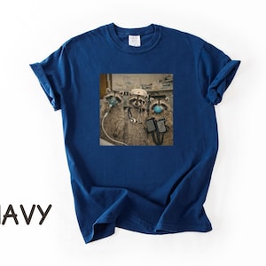 May include: Navy blue t-shirt featuring a square graphic of three raccoons wearing medical masks and equipment. The raccoons are depicted in a medical setting. The word "NAVY" is printed in large, brown letters at the bottom of the shirt.
