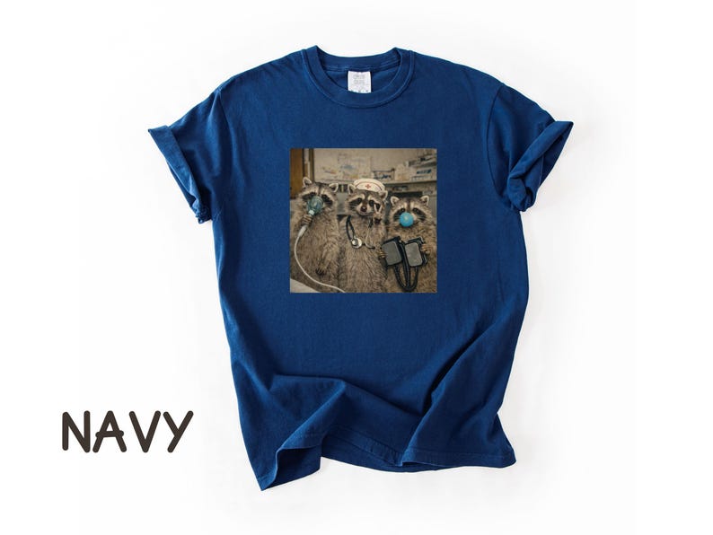 May include: Navy blue t-shirt featuring a square graphic of three raccoons wearing medical masks and equipment. The raccoons are depicted in a medical setting. The word "NAVY" is printed in large, brown letters at the bottom of the shirt.