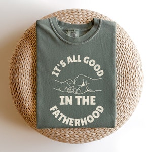 May include: A green t-shirt with the text "It's all good in the fatherhood" in white lettering. The text is surrounded by two fists bumping together.