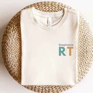 May include: A cream colored t-shirt with the text "Stephanie RT" in blue, green, and orange.