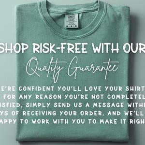 May include: A sage green t-shirt with the text "SHOP RISK-FREE WITH OUR Quality Guarantee". The shirt is folded neatly, with a tag visible at the collar. The text promises satisfaction.