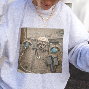 May include: A light gray sweatshirt featuring a graphic of three raccoons in medical attire. One raccoon wears a nurse's hat, another has an oxygen mask, and the third wears a blue surgical mask. The image has a vintage, slightly faded look.