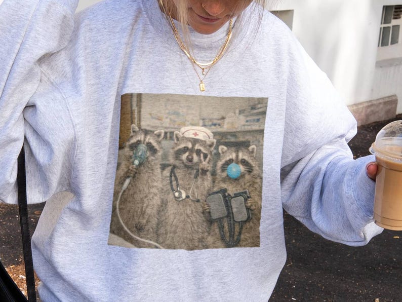 May include: A light gray sweatshirt featuring a graphic of three raccoons in medical attire. One raccoon wears a nurse's hat, another has an oxygen mask, and the third wears a blue surgical mask. The image has a vintage, slightly faded look.