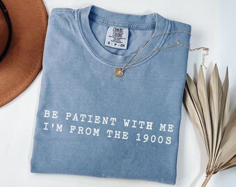 May include: A blue Comfort Colors t-shirt with the text "BE PATIENT WITH ME I'M FROM THE 1900S" in white. A gold necklace with a pendant is draped across the shirt. A brown hat and dried plant are in the background.