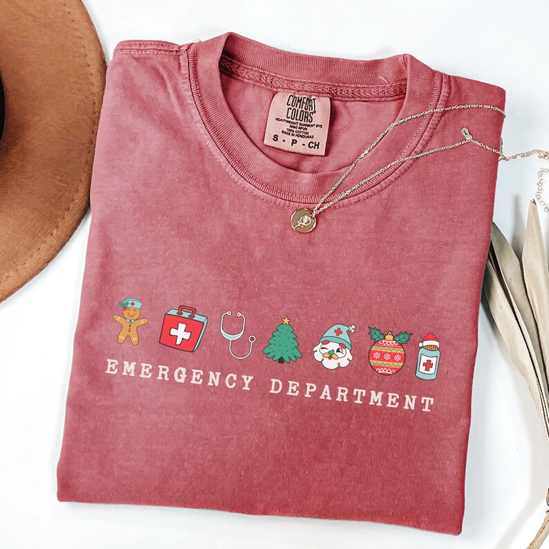 Peut inclure: Un t-shirt rouge avec un motif sur le th&egrave;me de No&euml;l repr&eacute;sentant un bonhomme en pain d'&eacute;pices, une trousse de premiers soins, un st&eacute;thoscope, un sapin de No&euml;l, le P&egrave;re No&euml;l, une boule de No&euml;l et une bouteille de m&eacute;dicaments. Le texte "EMERGENCY DEPARTMENT" est imprim&eacute; sous le motif.