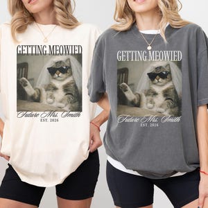 以下が含まれることがあります： クリーム色とチャコールグレーの2枚のTシャツ。それぞれサングラスとベールをつけた猫のグラフィックが特徴です。Tシャツのテキストは「GETTING MEOWIED」と「Future Mrs. Smith EST. 2026」です。