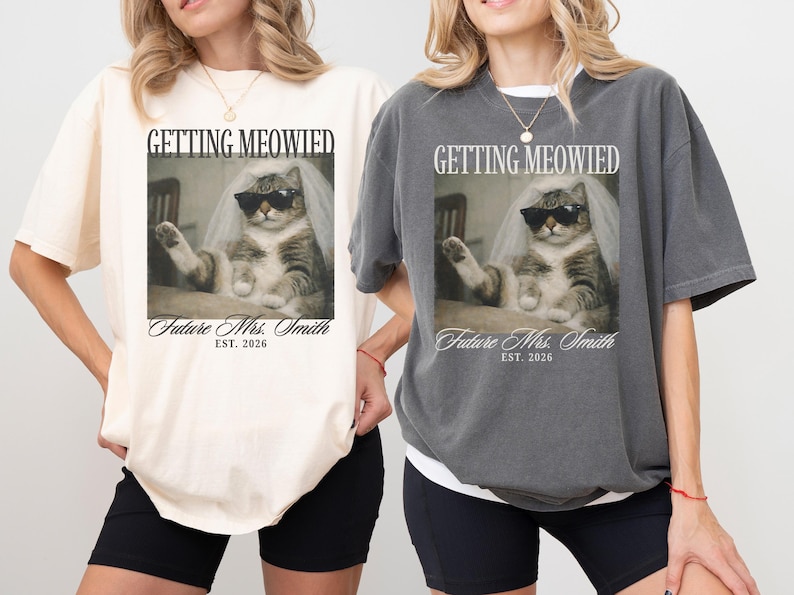 以下が含まれることがあります： クリーム色とチャコールグレーの2枚のTシャツ。それぞれサングラスとベールをつけた猫のグラフィックが特徴です。Tシャツのテキストは「GETTING MEOWIED」と「Future Mrs. Smith EST. 2026」です。