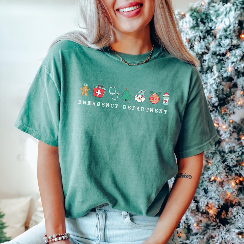 Peut inclure: Un t-shirt vert avec un motif sur le th&egrave;me de No&euml;l repr&eacute;sentant un bonhomme en pain d'&eacute;pices, une trousse de premiers soins, un st&eacute;thoscope, un sapin de No&euml;l, le P&egrave;re No&euml;l, une boule de No&euml;l et une bouteille de m&eacute;dicaments. Le texte "Emergency Department" est imprim&eacute; sous le motif.