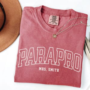 May include: A red t-shirt with the text "PARAPRO MRS. SMITH" in white. The shirt has a distressed look and is made of a soft, comfortable fabric.