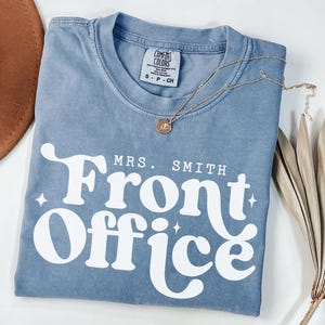 May include: A blue Comfort Colors t-shirt with the words "MRS. SMITH Front Office" in white, stylized font. A gold necklace with a pendant is draped on the shirt. A brown hat and dried palm leaves are visible.