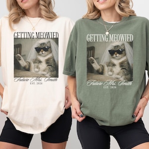 以下が含まれることがあります： クリーム色とオリーブグリーンの2枚のTシャツ。それぞれサングラスとベールをつけた猫が描かれています。「GETTING MEOWIED」と「Future Mrs. Smith EST. 2026」の文字が前面にプリントされています。