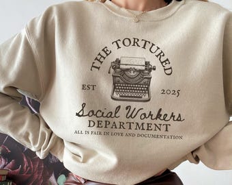 Puede incluir: Sudadera beige con un gráfico de una máquina de escribir vintage y el texto "THE TORTURED EST 2025 Social Workers DEPARTMENT ALL IS FAIR IN LOVE AND DOCUMENTATION"