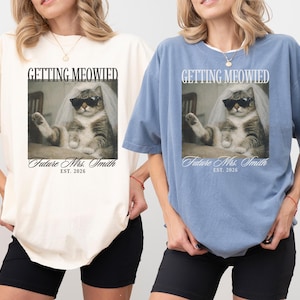 以下が含まれることがあります： クリーム色と青色の2枚のオーバーサイズのTシャツ。それぞれサングラスとベールをつけた猫が描かれています。「GETTING MEOWIED」と「Future Mrs. Smith EST. 2026」の文字が前面にプリントされています。黒いショートパンツと合わせています。
