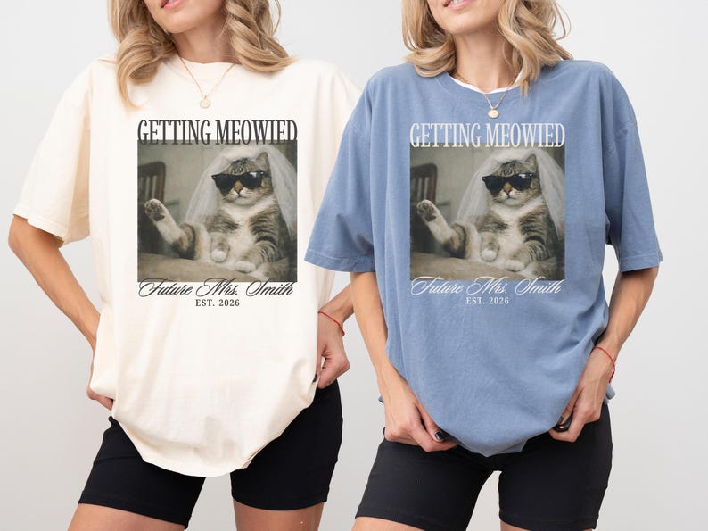 以下が含まれることがあります： クリーム色と青色の2枚のオーバーサイズのTシャツ。それぞれサングラスとベールをつけた猫が描かれています。「GETTING MEOWIED」と「Future Mrs. Smith EST. 2026」の文字が前面にプリントされています。黒いショートパンツと合わせています。