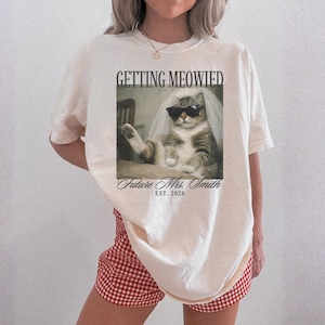以下が含まれることがあります： サングラスとベールをつけた猫のユーモラスな画像がプリントされたクリーム色のTシャツ。「GETTING MEOWIED」の文字が猫の上に、「Future Mrs. Smith EST. 2026」の文字が下にあります。 赤と白のチェック柄のショートパンツを着用しています。