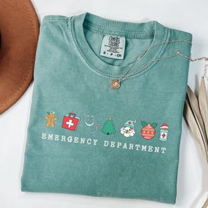 Peut inclure: Un t-shirt vert sarcelle avec un motif sur le th&egrave;me de No&euml;l qui dit "Service des urgences". Le motif repr&eacute;sente un bonhomme en pain d'&eacute;pices, une trousse de premiers soins, un st&eacute;thoscope, un sapin de No&euml;l, le P&egrave;re No&euml;l, une boule de No&euml;l et une bouteille de m&eacute;dicaments.