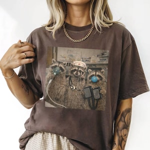 May include: Brown t-shirt featuring a humorous image of three raccoons dressed as medical professionals. One raccoon wears a nurse's hat, another has a blue bubble, and all have medical equipment. The shirt is a relaxed fit.