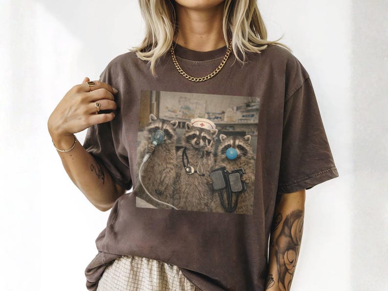 May include: Brown t-shirt featuring a humorous image of three raccoons dressed as medical professionals. One raccoon wears a nurse's hat, another has a blue bubble, and all have medical equipment. The shirt is a relaxed fit.