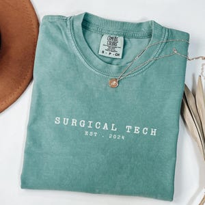 May include: A teal blue t-shirt with the text "SURGICAL TECH EST. 2024" in white lettering.