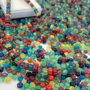 May include: A collection of colorful glass beads in various shapes and sizes. The beads are primarily in shades of red, green, blue, yellow, and purple. Some beads are strung together, while others are scattered around a white box, creating a vibrant display.