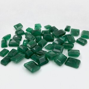 May include: A pile of emerald green gemstones, cut into rectangular shapes, with a polished finish. The gemstones are scattered on a white surface.