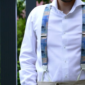 May include: Blue and white patterned suspenders with silver-tone metal clasps. The suspenders feature a scenic design with boats on a body of water. The suspenders are worn over a white button-down shirt.