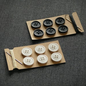 May include: Two cards with black and white buttons on them. The cards are on a gray surface. The black buttons are on a card with a needle and thread. The white buttons are on a card with a needle and thread.