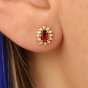 May include: Gold stud earrings featuring a red oval gemstone encircled by a halo of smaller clear gemstones.