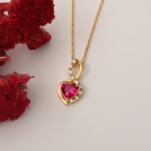 May include: A gold-colored necklace with a heart-shaped pendant. The pendant features a vibrant red gemstone surrounded by small, clear stones. A sprig of red flowers is in the background, set against a white backdrop.