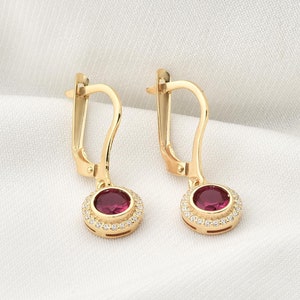 Könnte beinhalten: Ein Paar goldfarbene Ohrhänger. Jeder Ohrring hat einen runden, tiefroten Edelstein, der von kleineren, klaren Steinen umgeben ist. Die Ohrringe haben einen Klappverschluss.