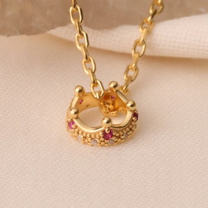 Op de afbeelding: Een gouden ketting met een klein kroontje als hanger. De kroon is versierd met kleine rode en witte steentjes.