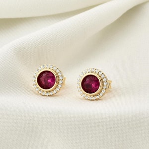 May include: A pair of gold earrings featuring a red gemstone set within a halo of smaller white gemstones.