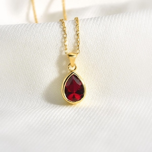 Op de afbeelding: Een goudkleurige ketting met een druppelvormige, dieprode edelsteen hanger. De hanger is gezet in een gouden bezel en hangt aan een fijne gouden ketting. De ketting wordt gepresenteerd op een witte, getextureerde stof.