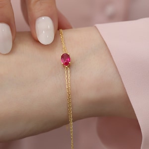 Könnte beinhalten: Ein zartes Armband aus einer goldenen Kette mit einem einzigen ovalen roten Edelstein.