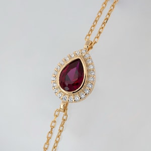 May include: A gold chain necklace with a pear-shaped red gemstone pendant surrounded by a halo of smaller white gemstones.