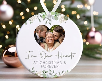 May include: White round Christmas bauble with a green floral design and a heart-shaped photo frame. The photo shows a woman with two boys. The bauble reads "In Our Hearts At Christmas & Forever".