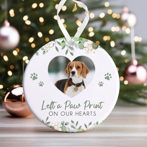 Puede incluir: Adorno navideño de cerámica blanca con un diseño floral verde y un marco de fotos en forma de corazón con un perro. El adorno dice "Left a Paw Print on Our Hearts".