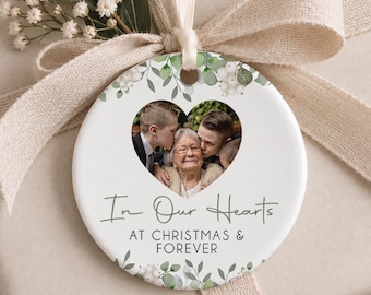 May include: A round white ceramic ornament featuring a heart-shaped photo of an older person being kissed by two youths. The text reads In Our Hearts AT CHRISTMAS & FOREVER. The ornament is decorated with green leaves and white berries and hangs by a ribbon.