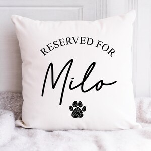 Puede incluir: Un coj&iacute;n cuadrado blanco con el texto "RESERVED FOR Milo" en escritura negra. Debajo del nombre hay una huella de pata negra. El coj&iacute;n est&aacute; sobre una superficie blanca.