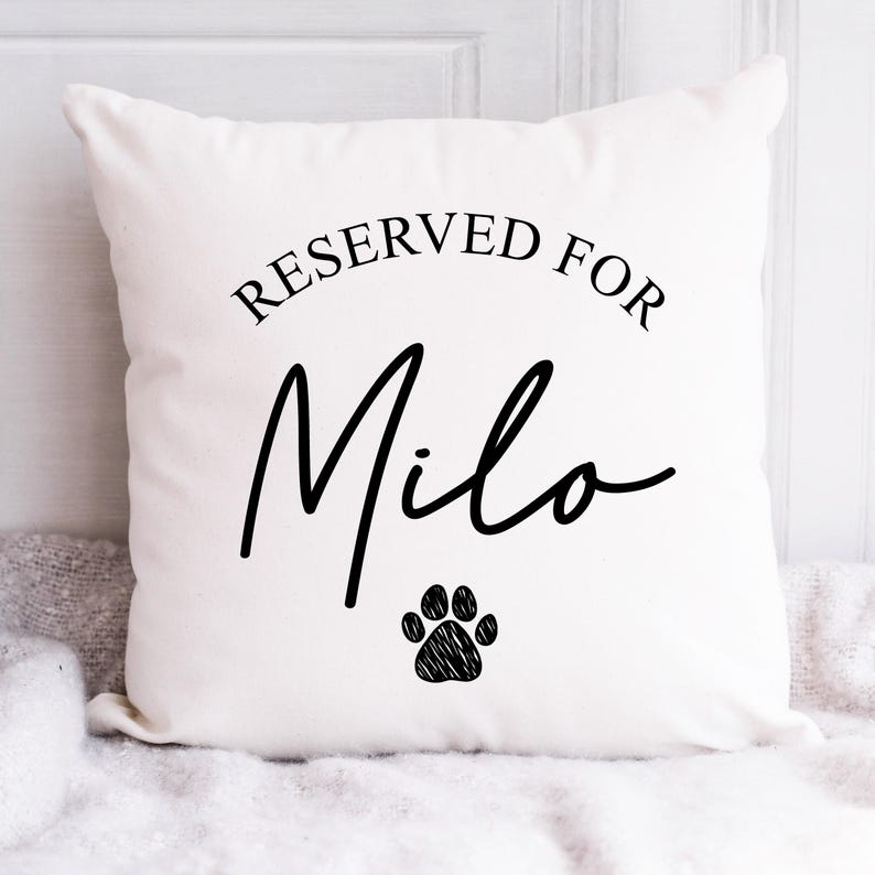 Puede incluir: Un coj&iacute;n cuadrado blanco con el texto "RESERVED FOR Milo" en escritura negra. Debajo del nombre hay una huella de pata negra. El coj&iacute;n est&aacute; sobre una superficie blanca.
