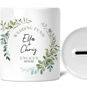 May include: White ceramic wedding fund money jar with a green leafy design and the text "Wedding Fund Ella + Chris Engaged 23/11/23".