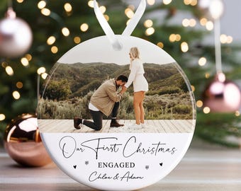 May include: A white round Christmas ornament with a photo of a couple getting engaged. The text on the ornament reads "Our First Christmas Engaged Chloe & Adam".